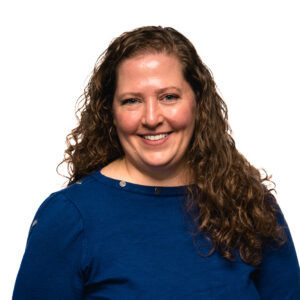 Bess Jayme, with long curly brown hair smiles directly at the viewer while wearing a solid dark blue long-sleeved shirt.