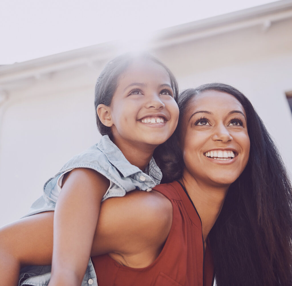 A smiling woman with long dark hair carries a young girl on her back against a bright, softly blurred building exterior.