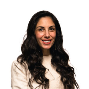 Holly Stephens, with long, dark, wavy hair smiles warmly at the camera against a plain white background, wearing a beige top.