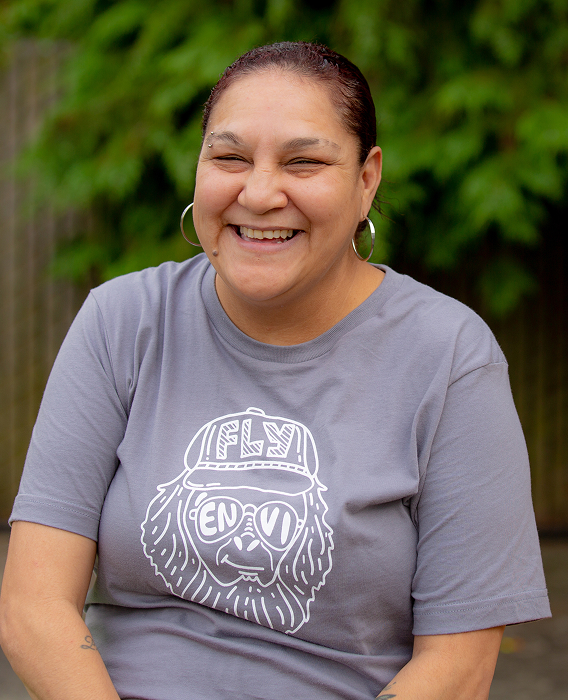 A smiling person with dark hair pulled back wears a grey t-shirt and hoop earrings.