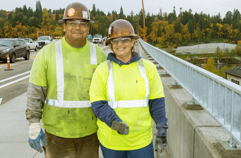 Two construction workers in high-visibility vests and hard hats stand smiling on a concrete bridge over a tree-lined area.