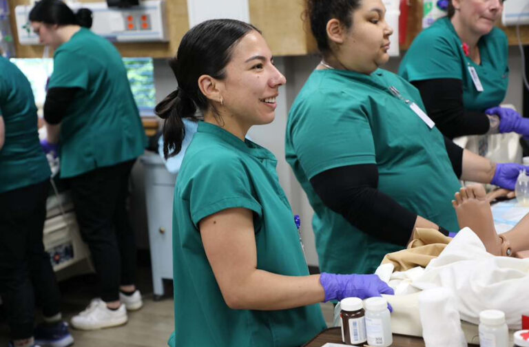 Two healthcare workers in green scrubs and purple gloves provide care to a patient while standing in a busy workspace.