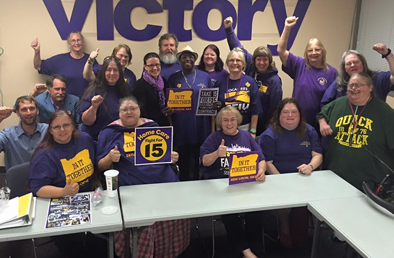 A diverse group of people stand in a large meeting room and pose together in front of a wall that has the word VICTORY.