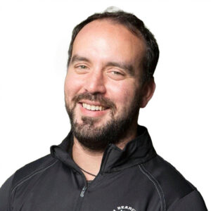 Kosti Efstathiou, with a dark beard and short brown hair smiles warmly at the camera while wearing a black athletic zip-up jacket.