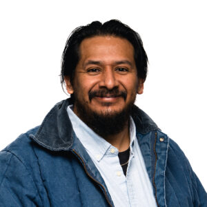 Miguel Rodriguez with dark hair and a beard smiles warmly, wearing a blue jacket over a light button-up shirt.