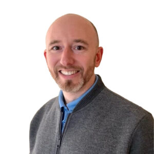 Nathan Biel, a bald man with a short, trimmed beard smiles at the camera, wearing a light blue polo shirt under a grey zip-up jacket.