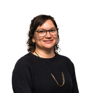 Dark-haired Sara Duerr with shoulder-length curls smiles at the camera while wearing thin-rimmed glasses and a black sweater.