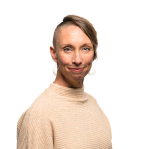 A person with a short, undercut hairstyle smiles gently at the camera while wearing a textured beige high-neck sweater.