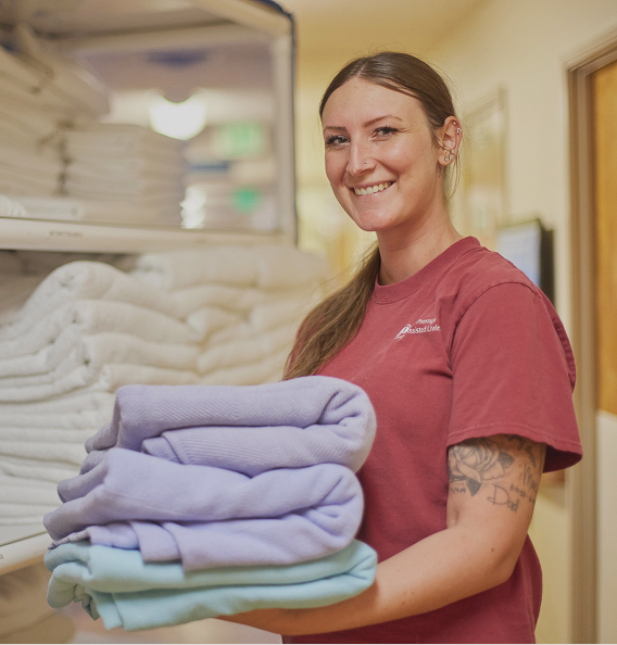 A smiling long haired in a dark red t-shirt holds several folded pastel towels while standing near a large supply closet.
