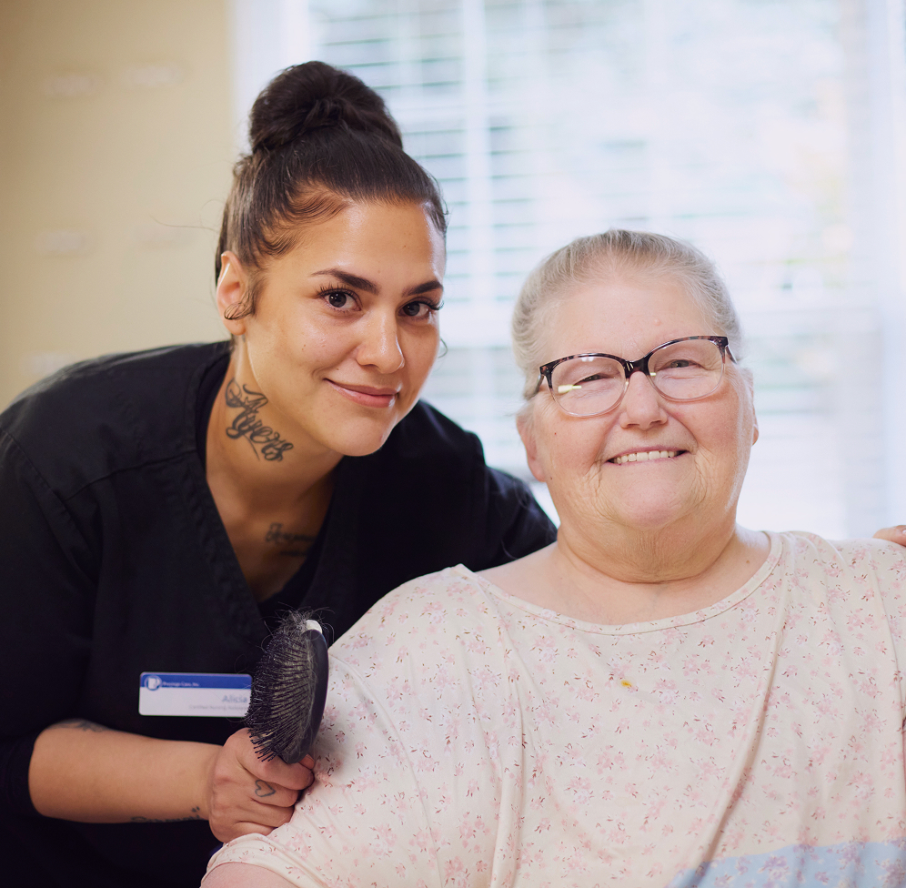 An older woman with glasses smiles brightly, seated in front of a caregiver who holds a hairbrush near her shoulder.