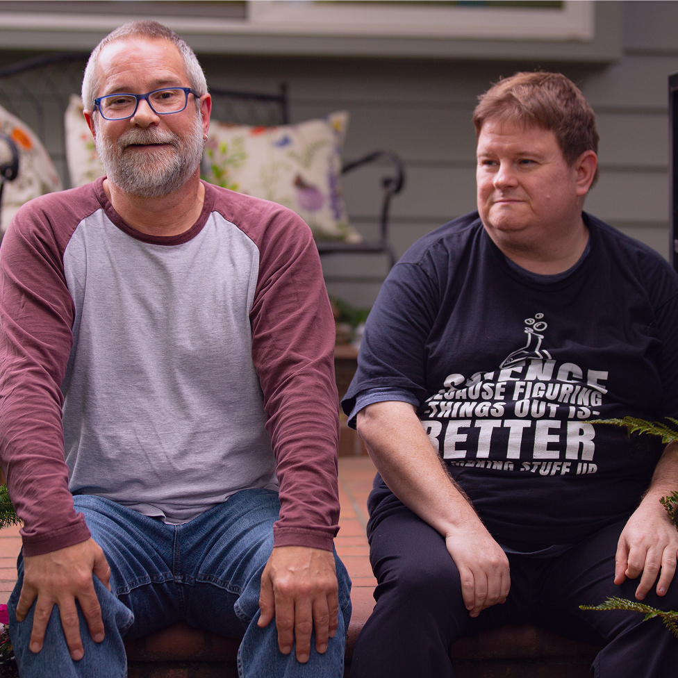 A person wearing a black shirt with text about science sits on a porch bench beside another person with a gray long-sleeve.