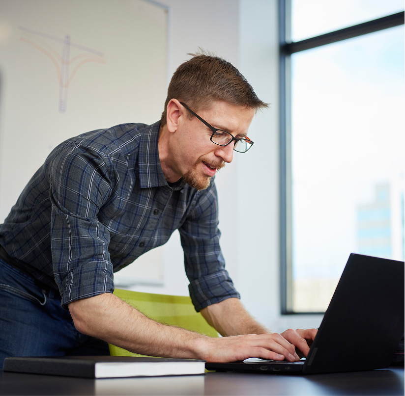A man in a plaid shirt and glasses leans over a desk, typing on his laptop while looking intensely at the glowing screen.