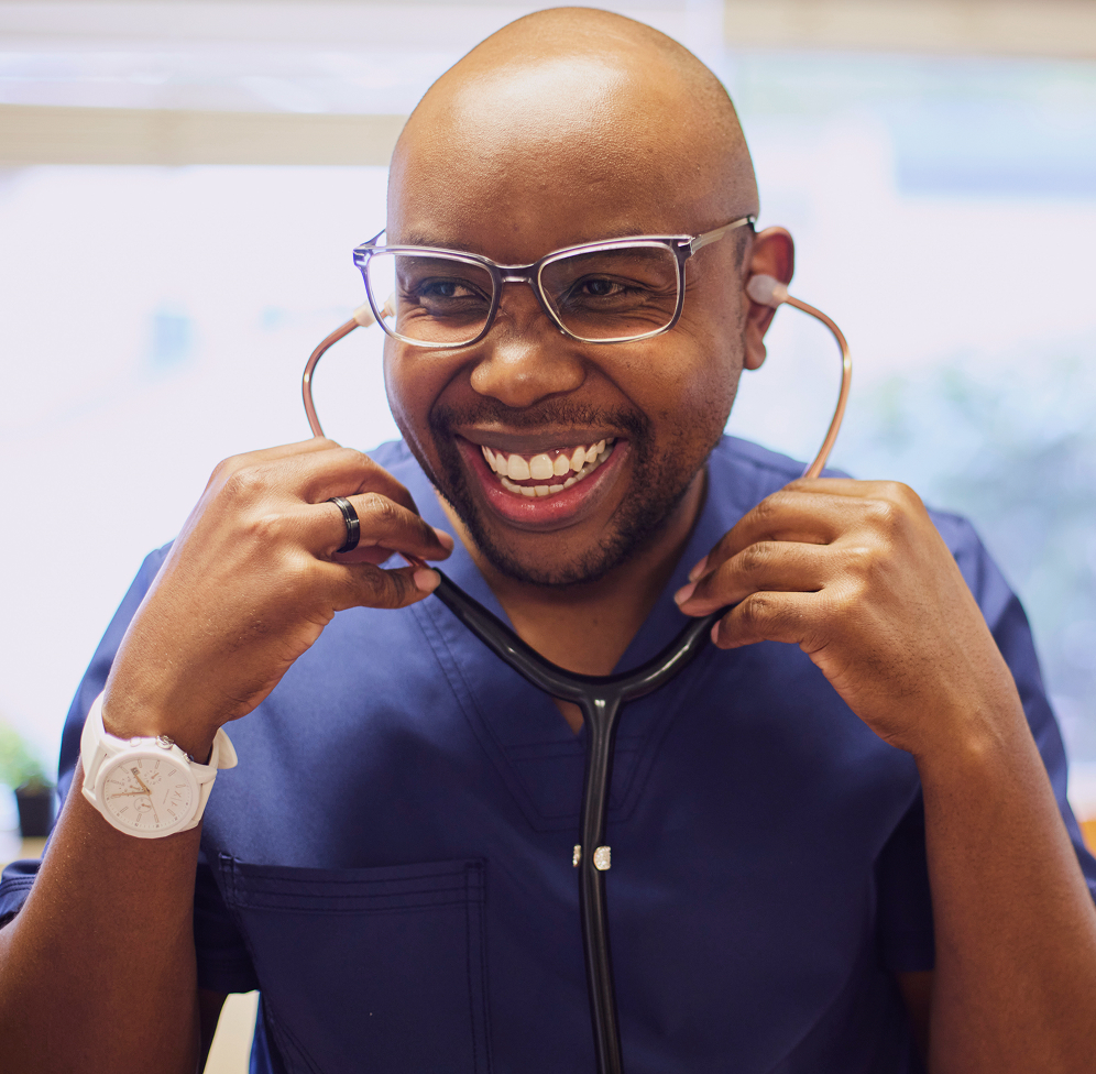 A smiling medical professional with a shaved head and glasses holds a stethoscope to their ears while wearing blue scrubs.