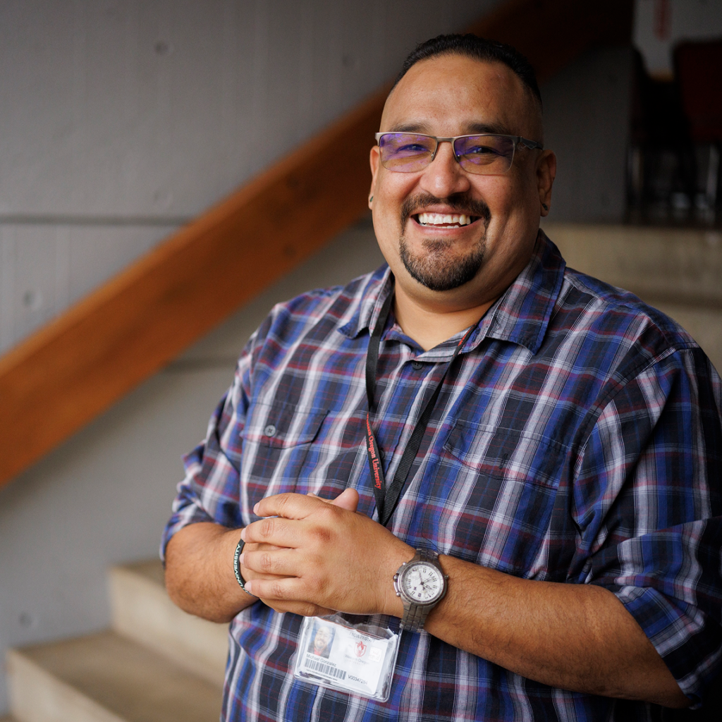 A smiling person with glasses, a goatee, and a plaid shirt stands with hands clasped in front on a staircase.