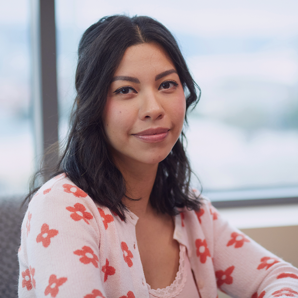 A woman with dark hair smiles gently while wearing a light pink cardigan decorated with a repeating pattern of red flowers.
