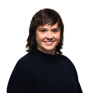 Ann Sacia, with short dark brown hair and bangs smiles warmly at camera, wearing a simple black knit sweater against white.