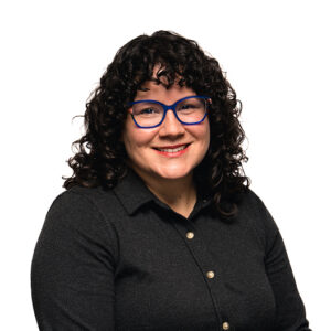 Katie Coombs, dark curly hair framing the face, smiles at the camera while wearing blue glasses and a grey polo shirt.