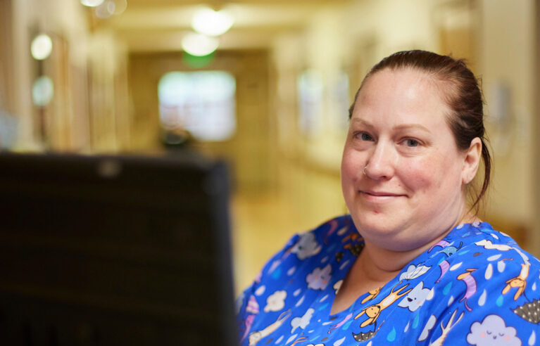 A woman in a blue scrub top patterned with clouds and dogs smiles while working at a computer screen.