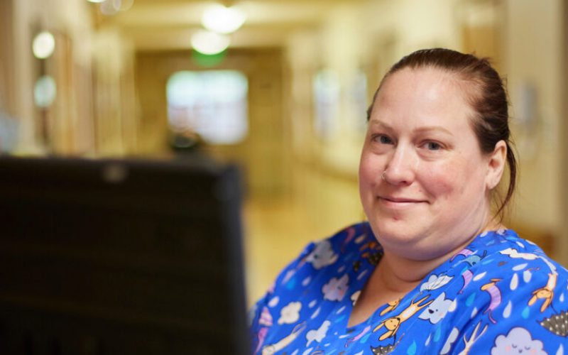 A woman in a blue scrub top patterned with clouds and dogs smiles while working at a computer screen.