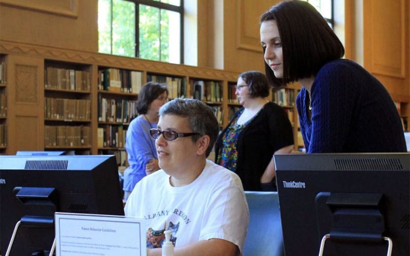 Two women working on a library computer, one standing and one sitting